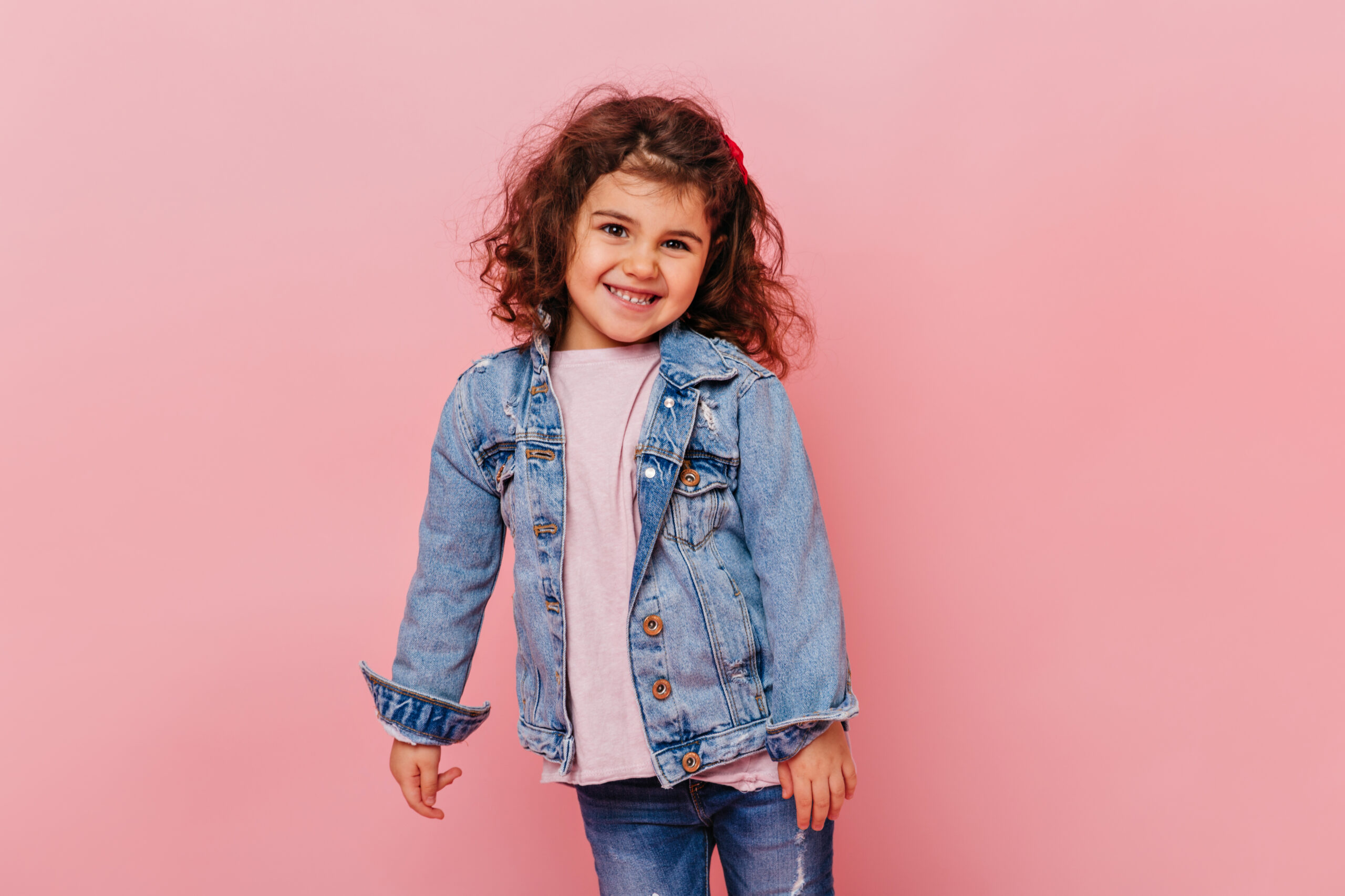 Smiling little girl with wavy hair standing on pink background. Studio shot of adorable preteen kid wearing denim jacket.